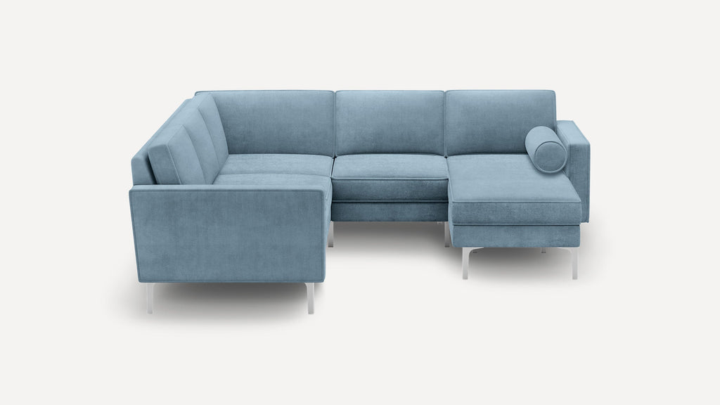 Modern blue velvet L-shaped sectional sofa with metal legs and a cylindrical bolster pillow on the chaise lounge side.