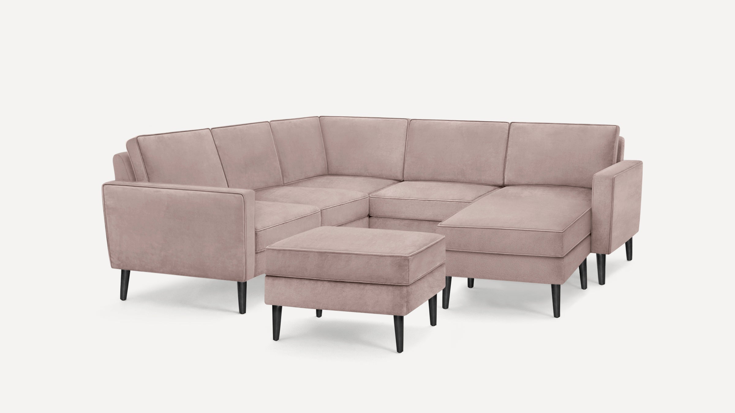 Modern blush pink L-shaped sectional sofa with matching ottoman, black tapered legs, and clean-lined cushions on white background.