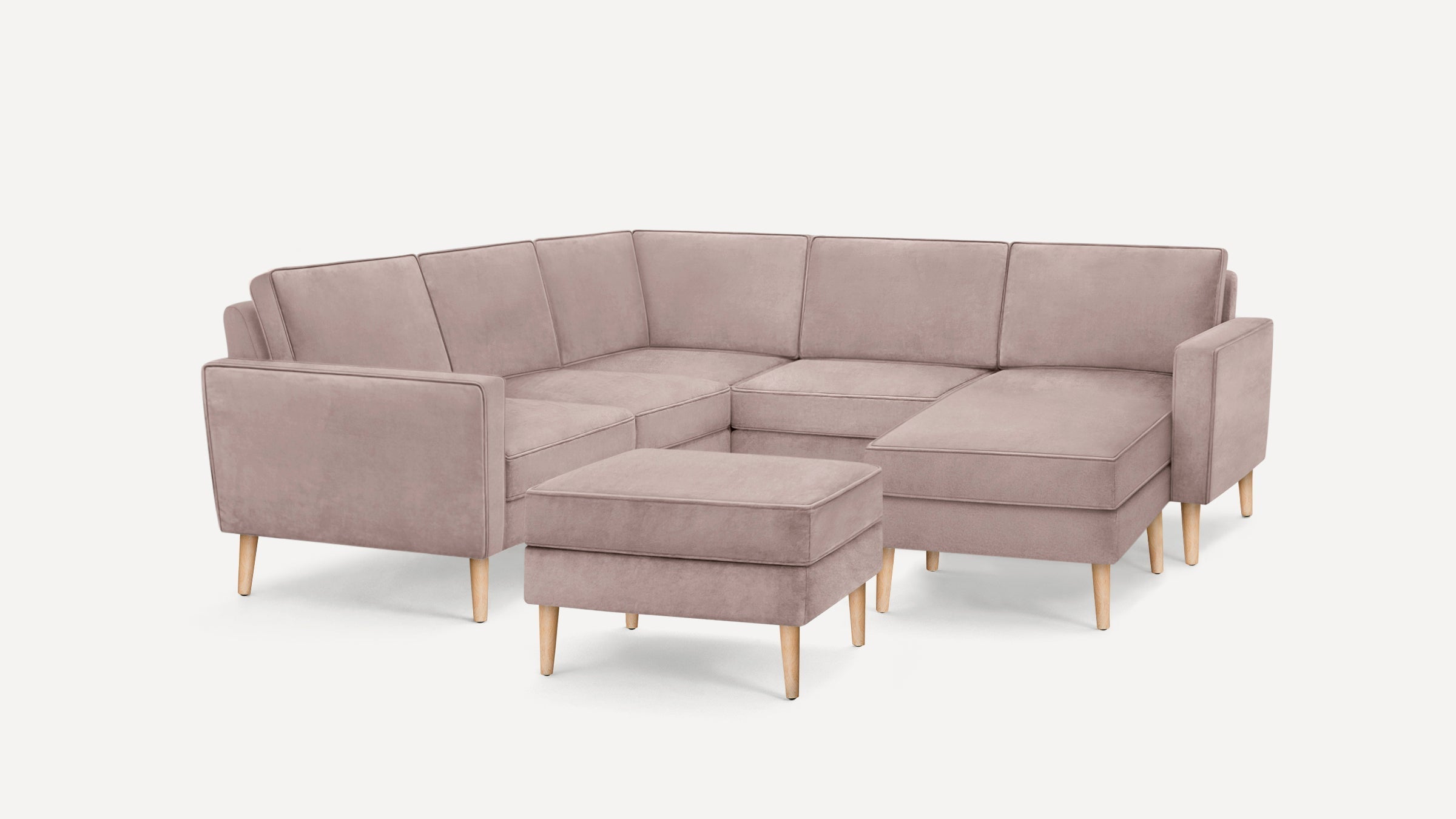Light mauve L-shaped sectional sofa with wooden legs and matching ottoman on a white background.