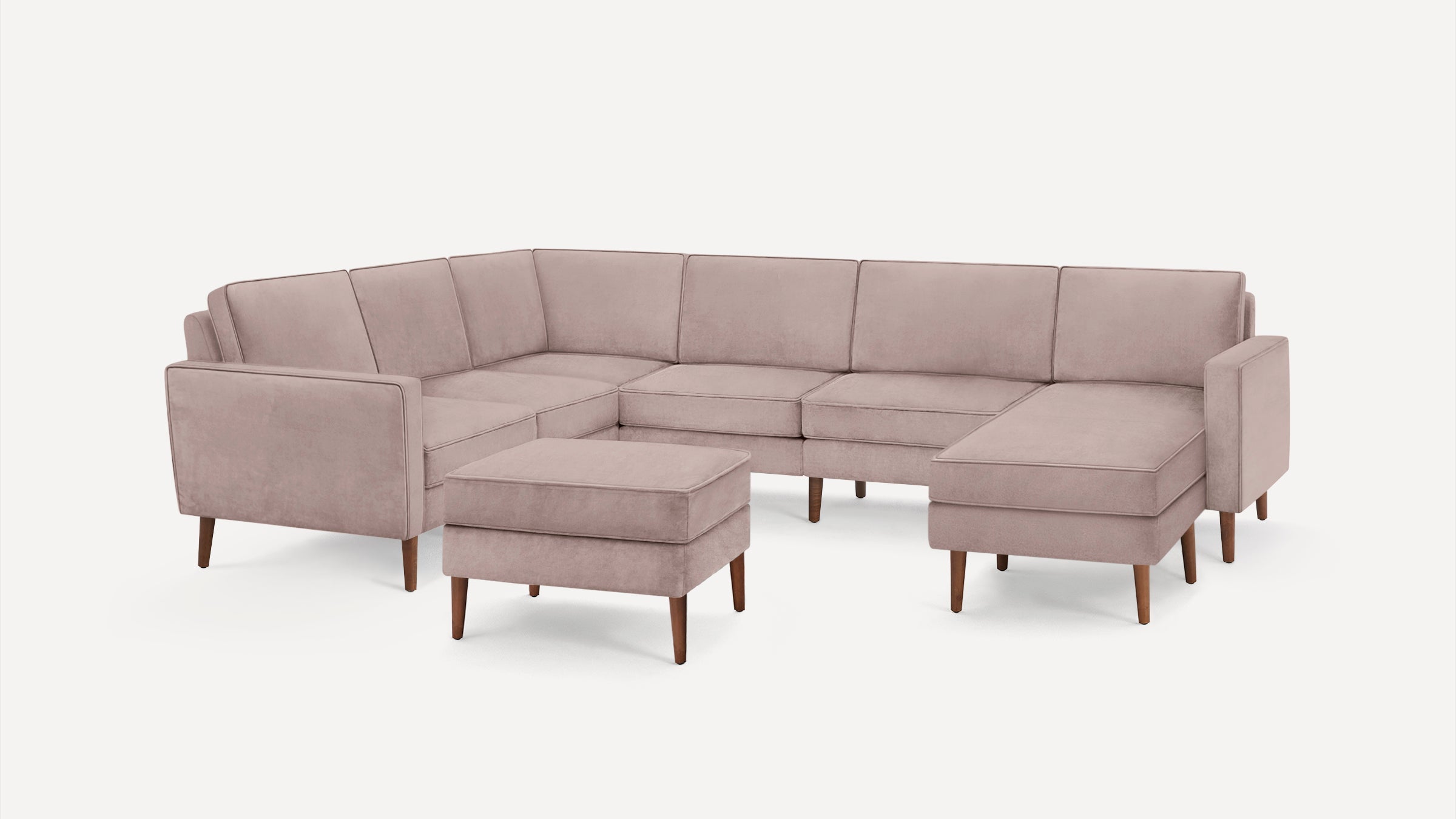 Light pink velvet U-shaped sectional sofa with wooden legs and matching ottoman, modern and minimalist design.