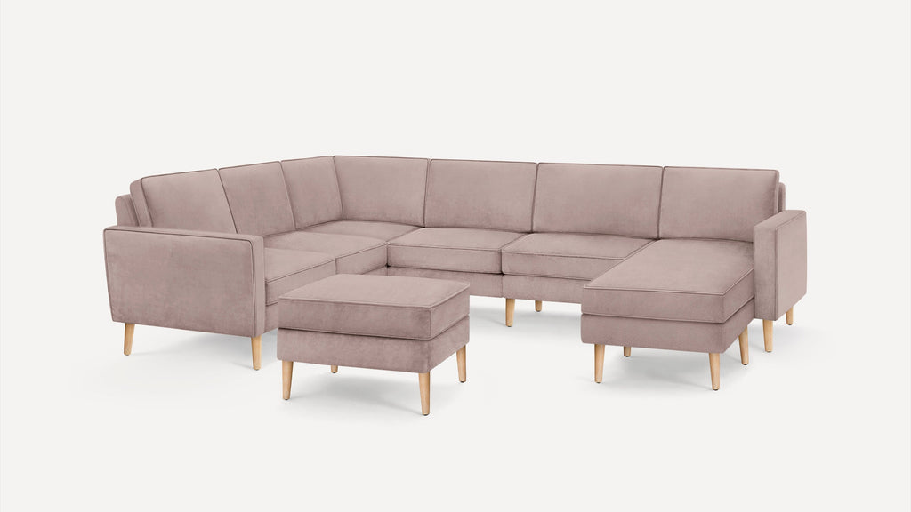 Light pink L-shaped sectional sofa with matching ottoman, featuring clean lines, cushioned seats, and light wooden tapered legs on white background.