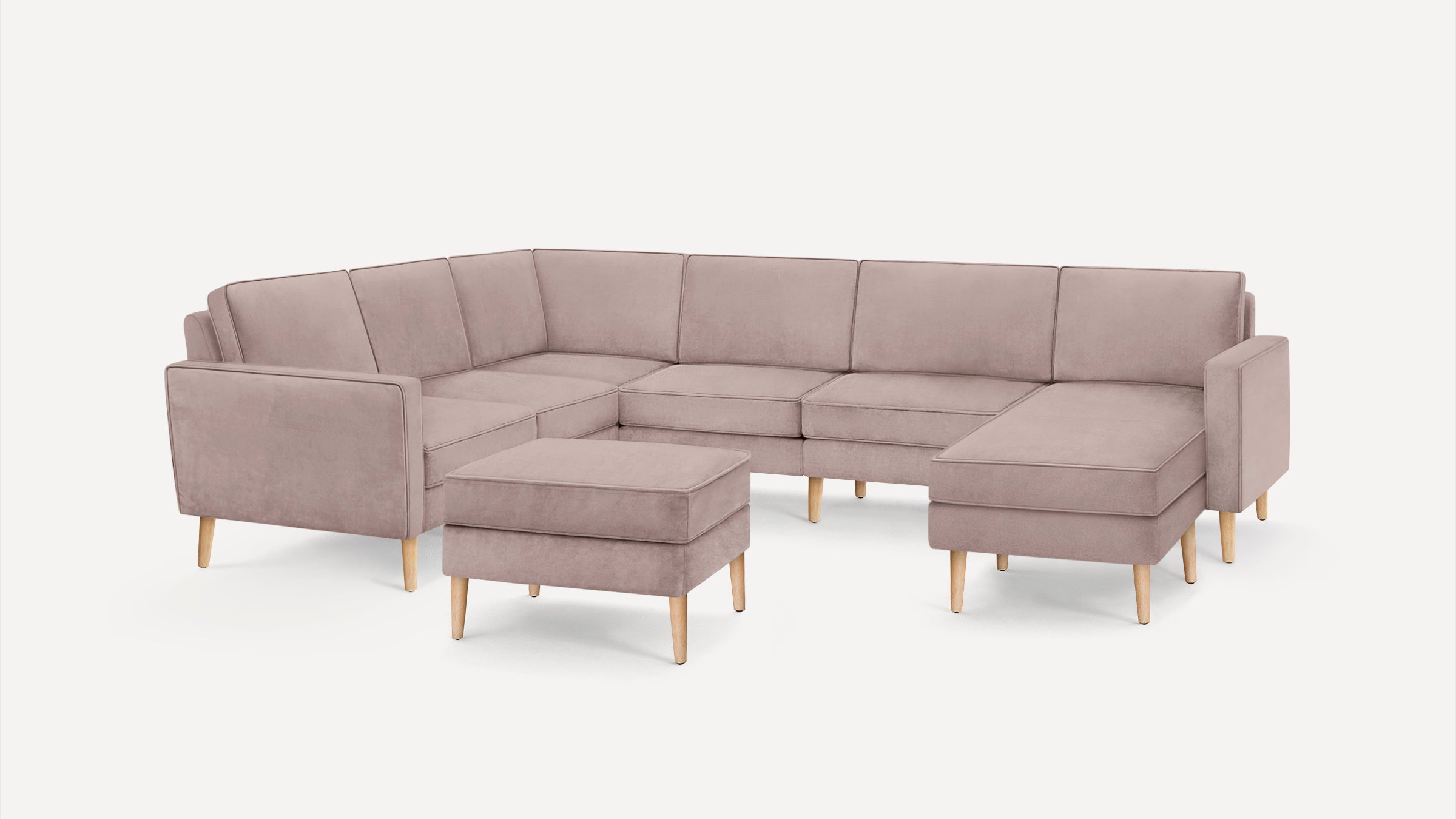 Light pink L-shaped sectional sofa with matching ottoman, featuring clean lines, cushioned seats, and light wooden tapered legs on white background.