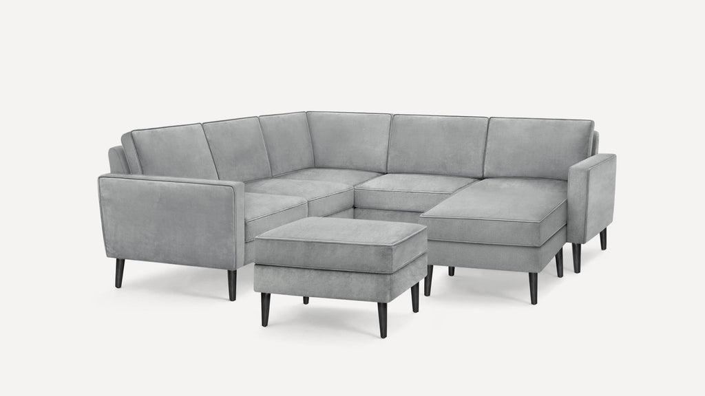 Modern gray velvet sectional sofa with black tapered legs and matching ottoman on white background.