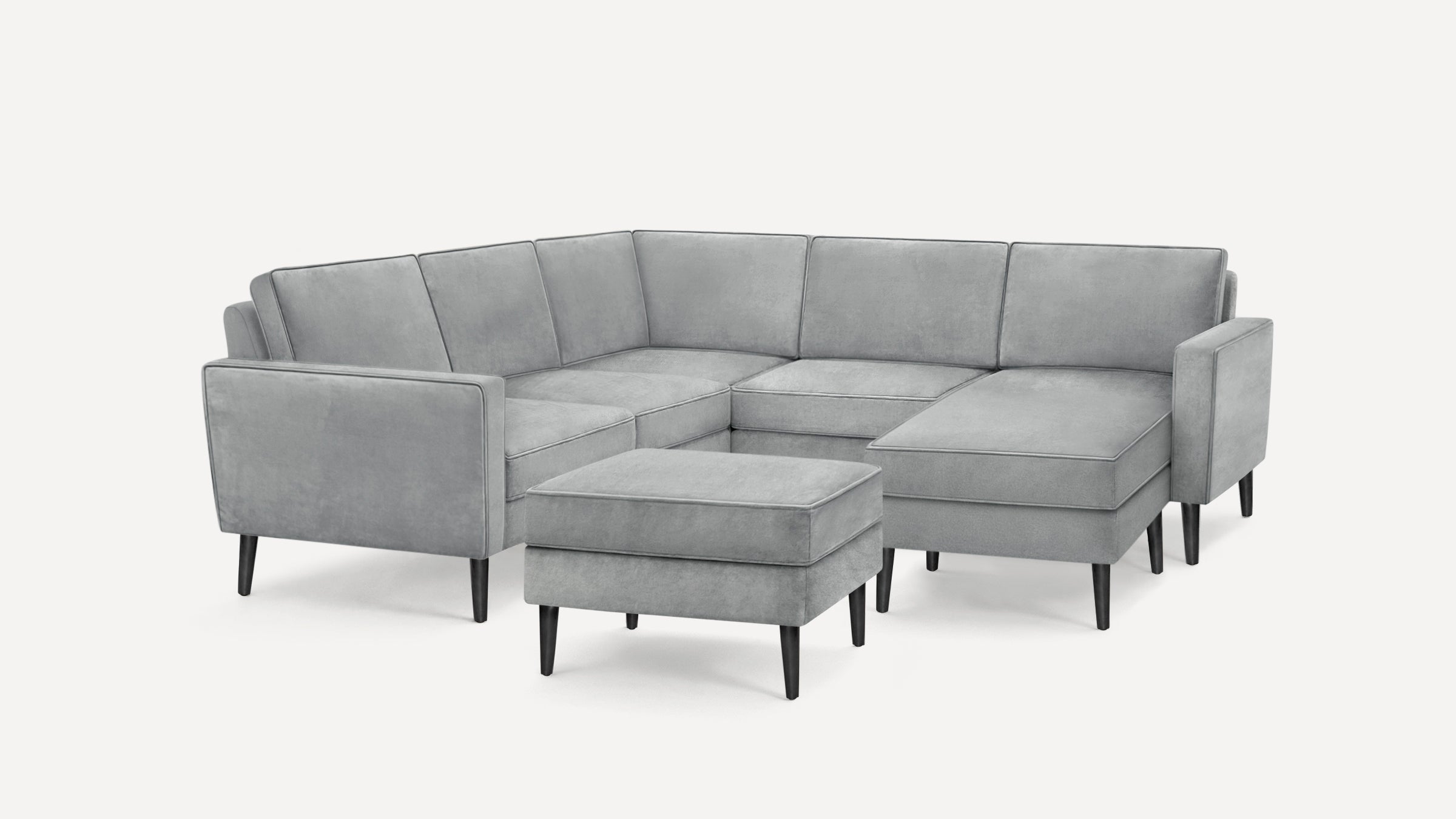 Modern gray velvet sectional sofa with black tapered legs and matching ottoman on white background.