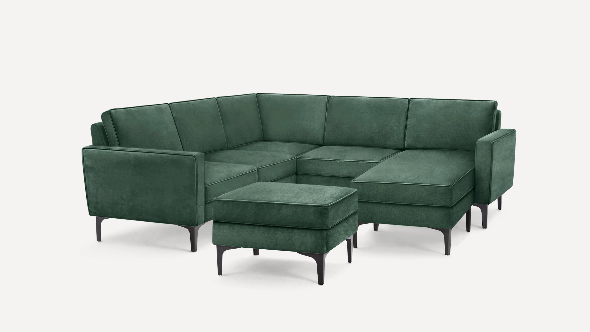 Modern dark green velvet L-shaped sectional sofa with matching ottoman and slim black legs on white background.