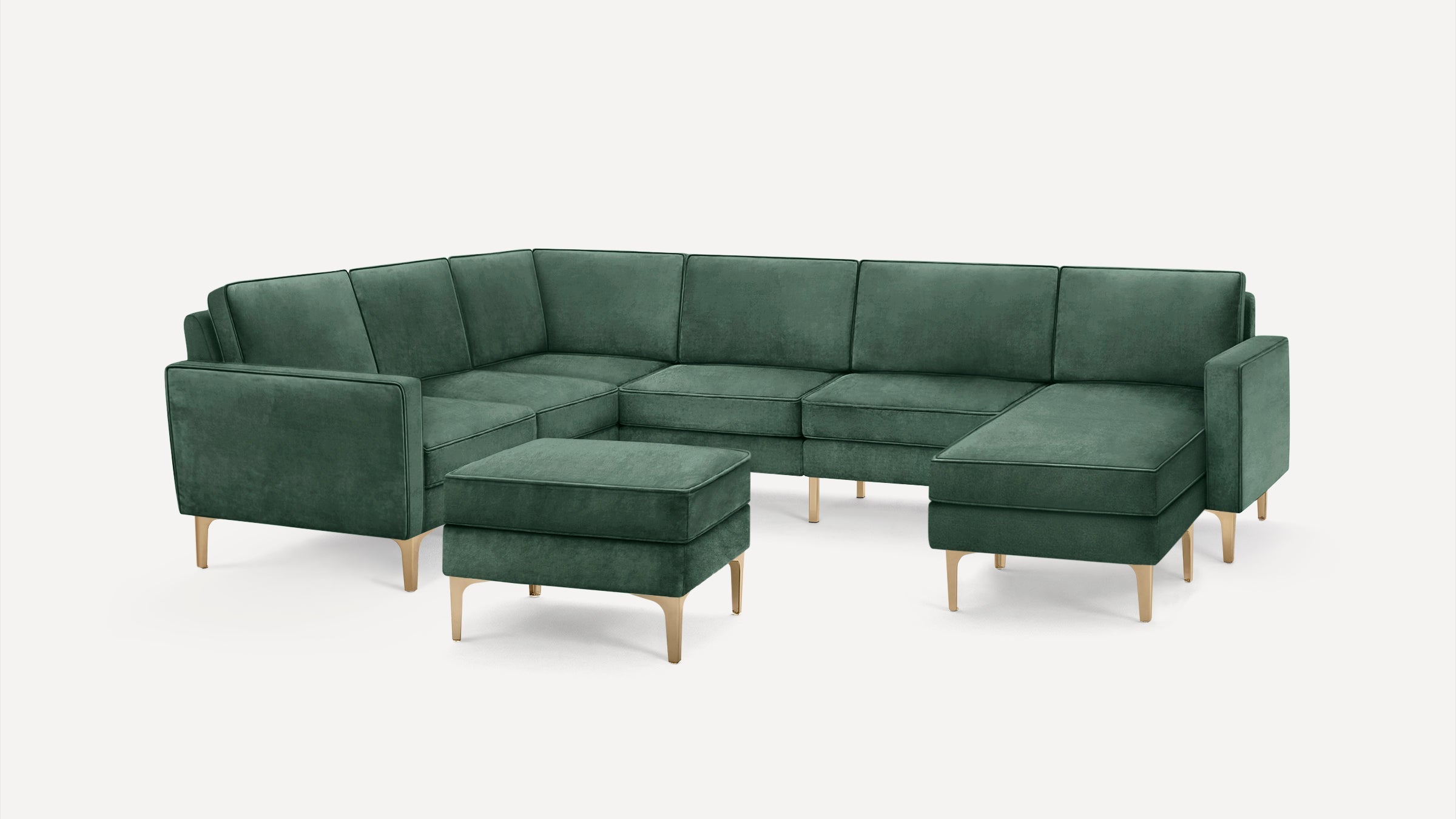 Green velvet U-shaped sectional sofa with brass legs and matching ottoman against white background.