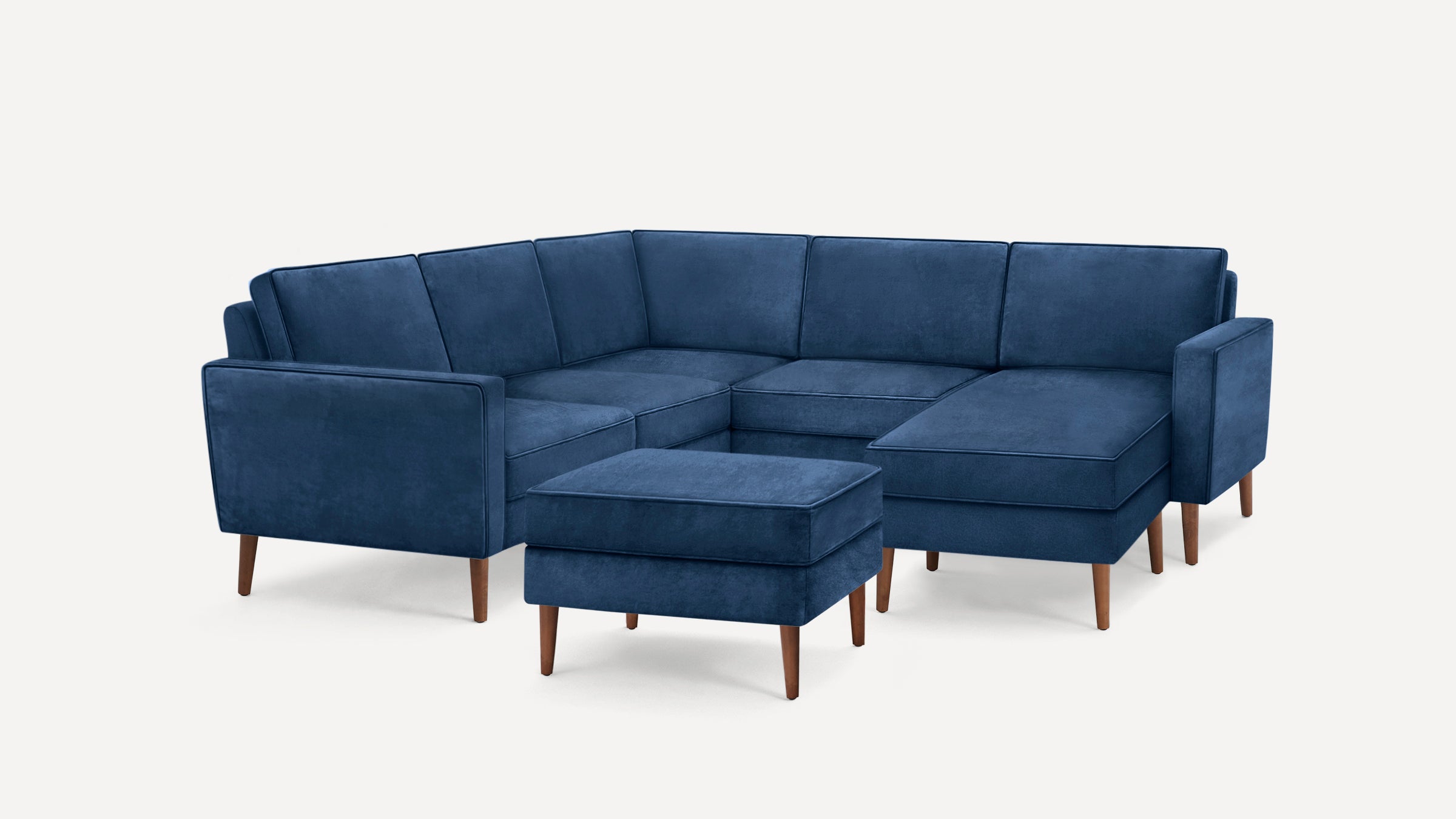 Modern navy blue velvet L-shaped sectional sofa with wooden legs and matching ottoman on white background.