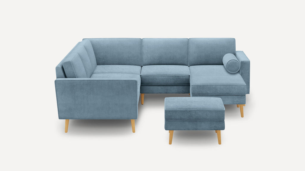 Modern blue L-shaped sectional sofa with wooden legs, a matching ottoman, and a cylindrical bolster pillow on chaise lounge.