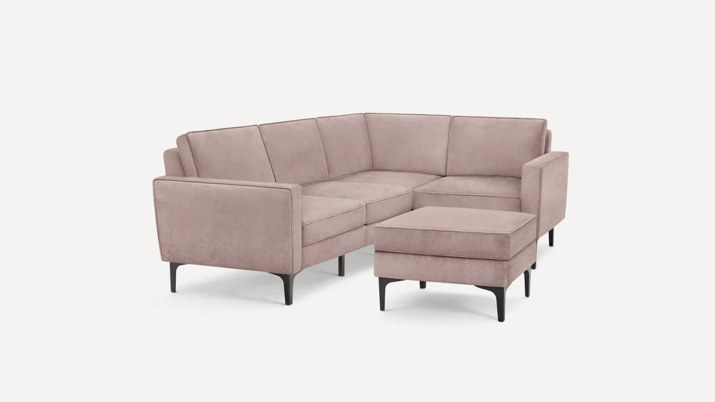 Modern L-shaped sectional sofa with blush pink velvet upholstery, clean lines, and black tapered legs, paired with a matching ottoman.