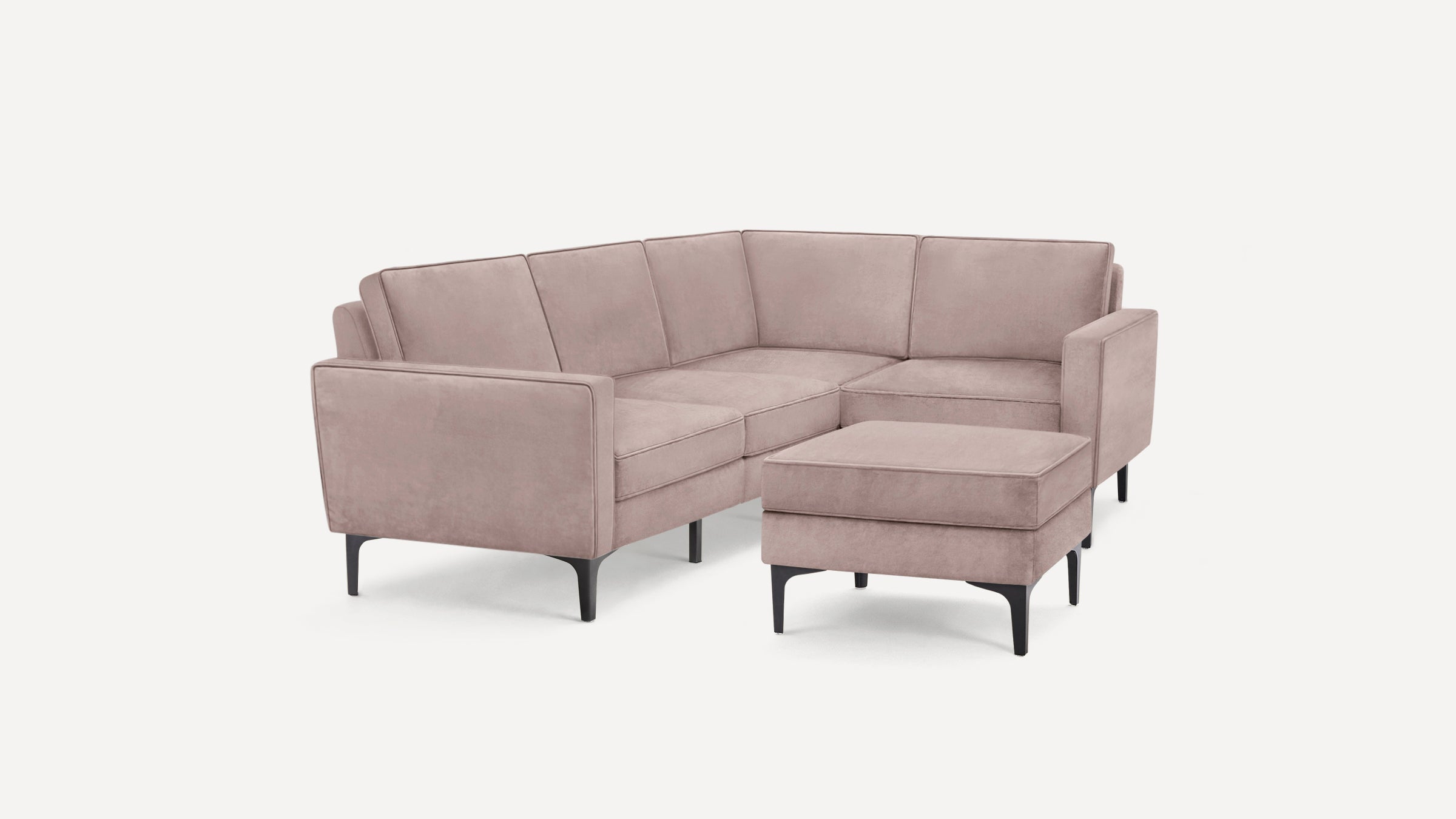 Modern L-shaped sectional sofa with blush pink velvet upholstery, clean lines, and black tapered legs, paired with a matching ottoman.