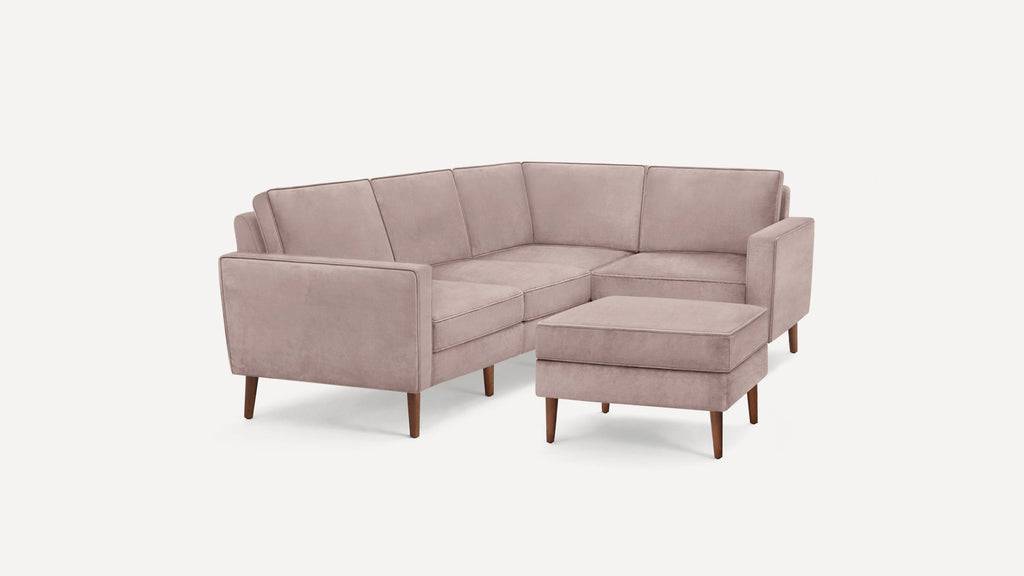 Light pink velvet L-shaped sectional sofa with wooden legs and matching ottoman on a white background.