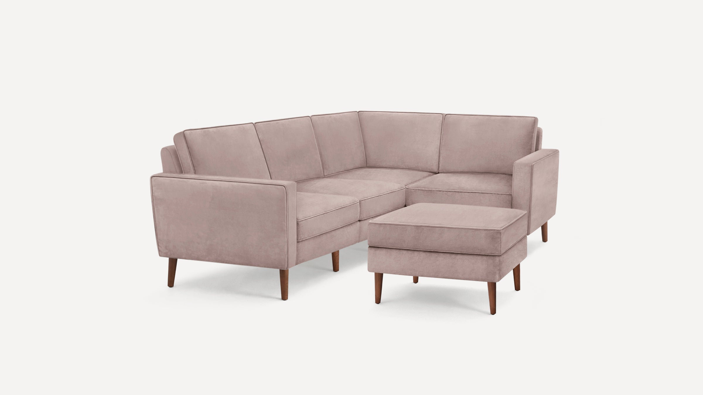 Light pink velvet L-shaped sectional sofa with wooden legs and matching ottoman on a white background.