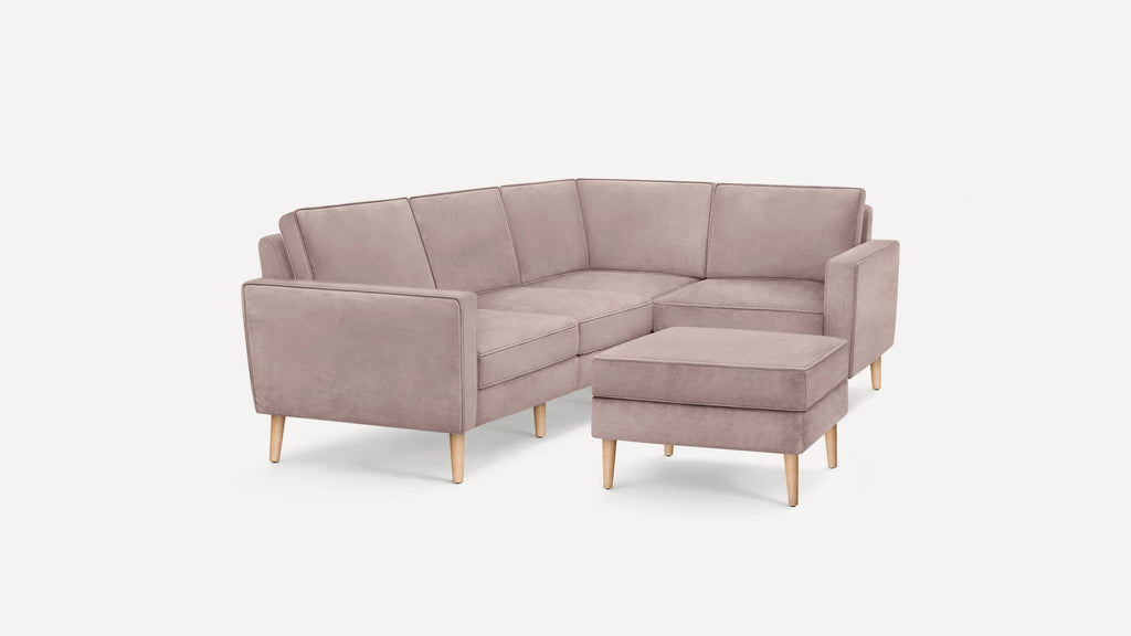 Light mauve L-shaped sectional sofa with wooden legs and matching ottoman on white background.