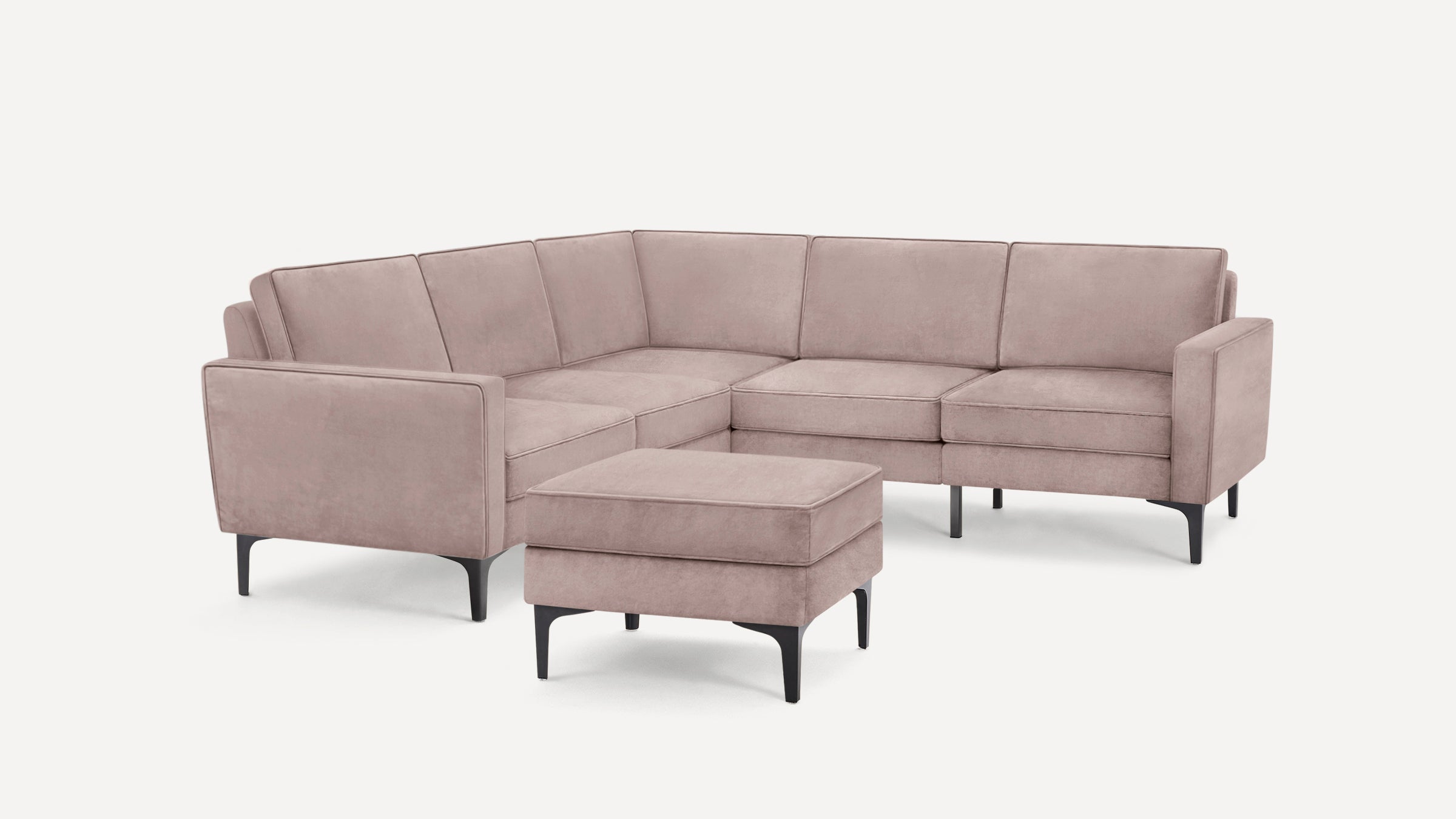 Modern L-shaped sectional sofa in soft dusty pink fabric with matching ottoman and black tapered legs on white background.
