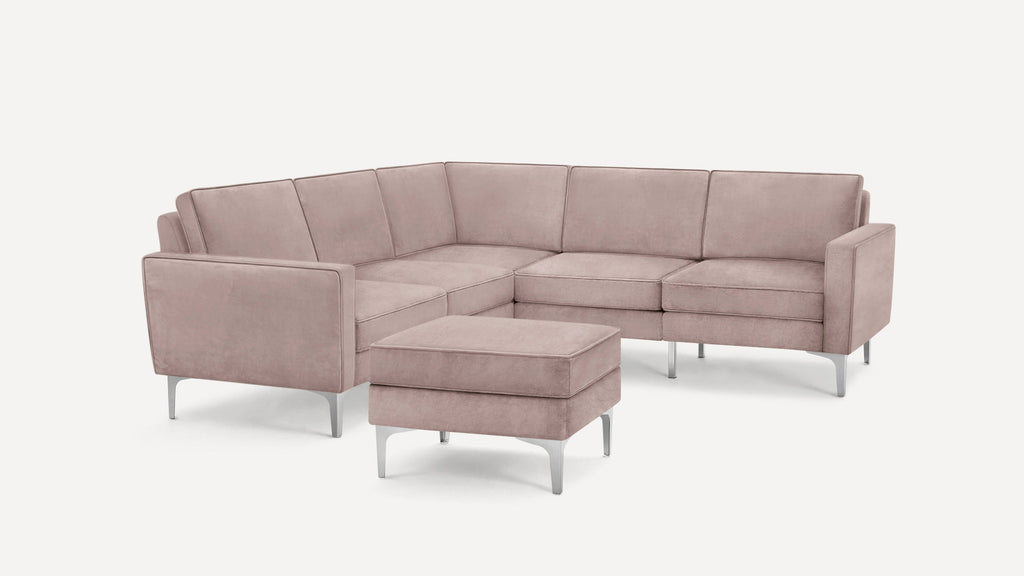 Light pink upholstered L-shaped sectional sofa with metal legs and matching ottoman on white background.