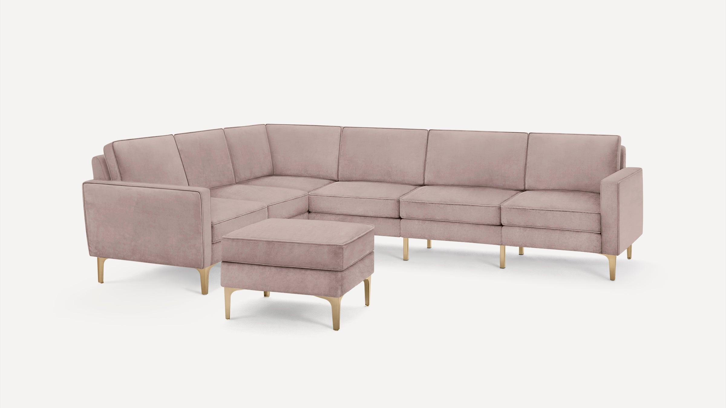 Modern blush pink velvet L-shaped sectional sofa with matching ottoman and brass-finished legs on white background.