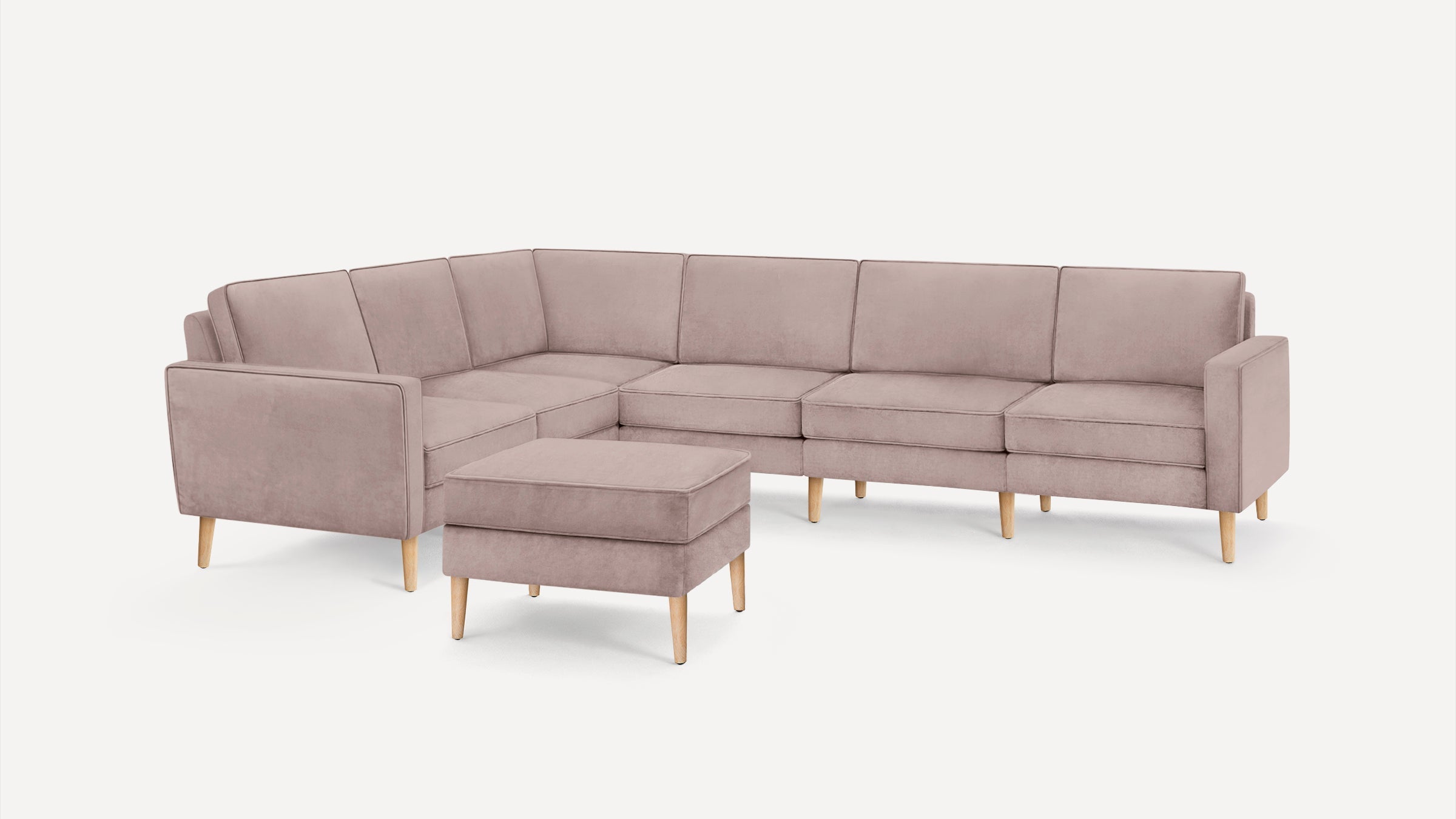 Light pink modern L-shaped sectional sofa with wooden legs and a matching cushioned ottoman on white background.