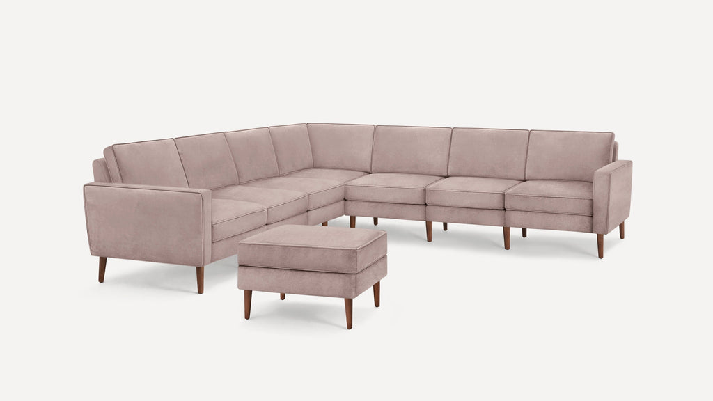 L-shaped dusty pink velvet sectional sofa with wooden legs and matching ottoman on white background.