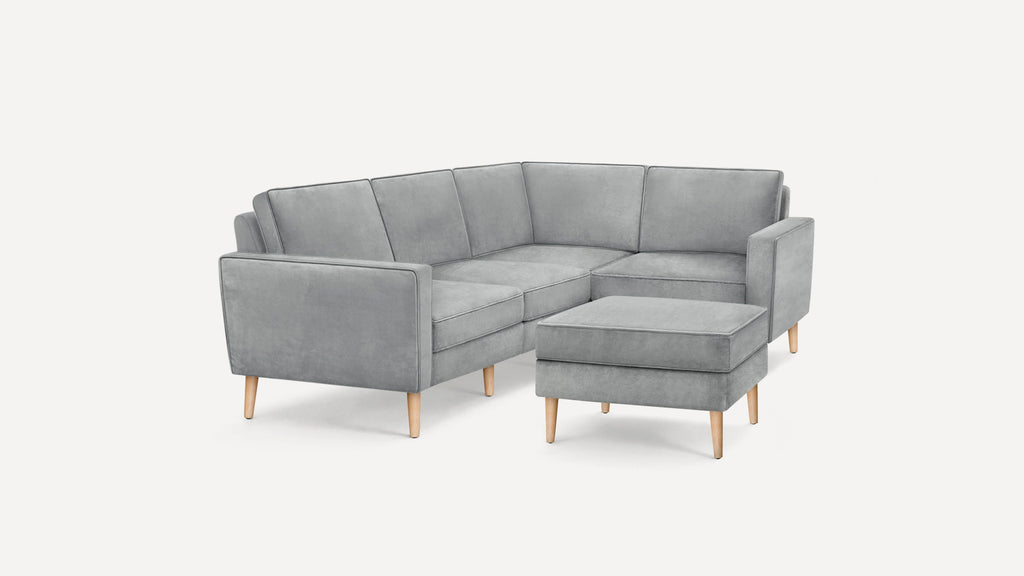Modern gray velvet L-shaped sectional sofa with matching ottoman, supported by light wooden tapered legs.