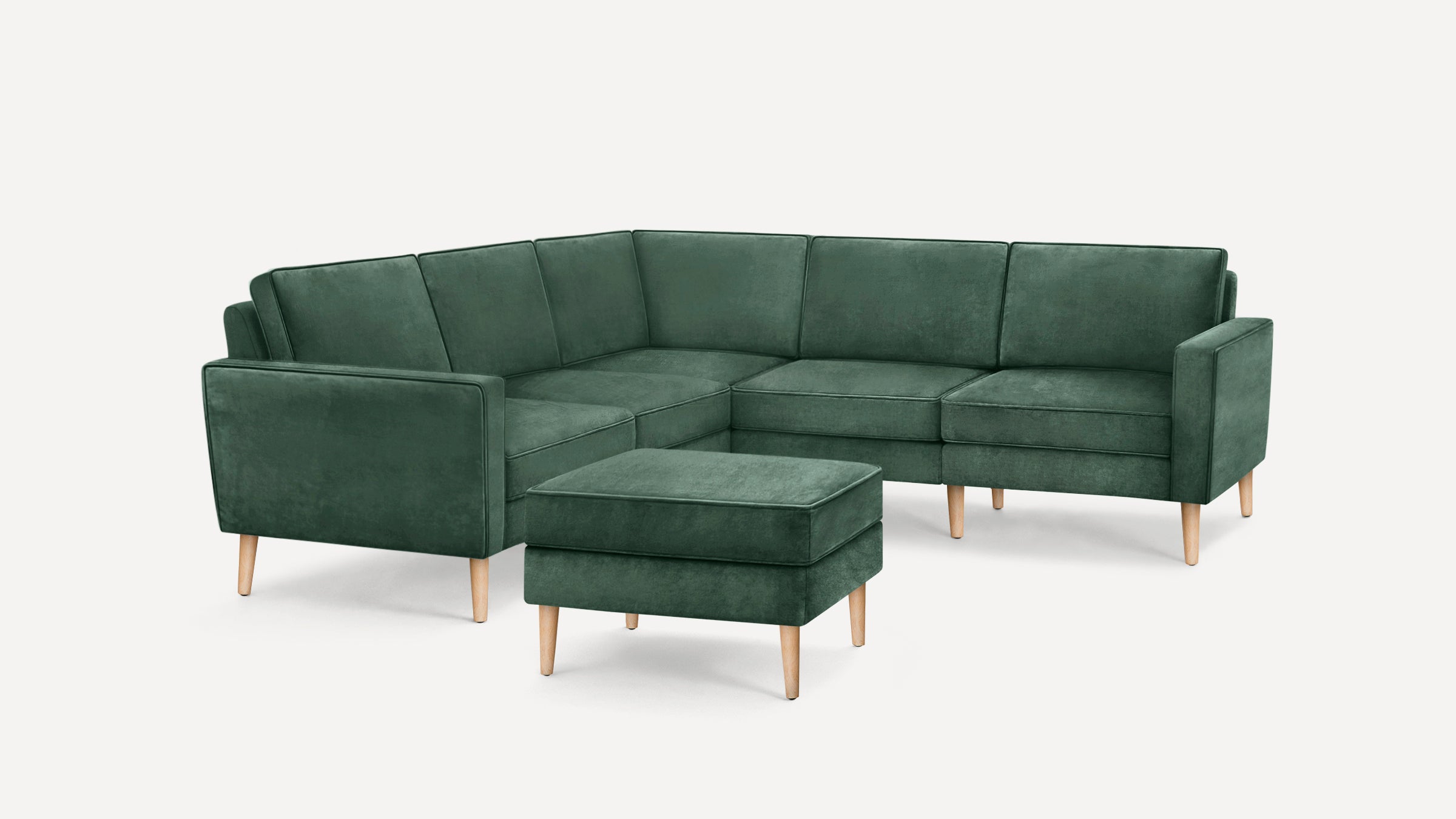 Dark green velvet L-shaped sectional sofa with light wooden legs and matching ottoman on a white background.