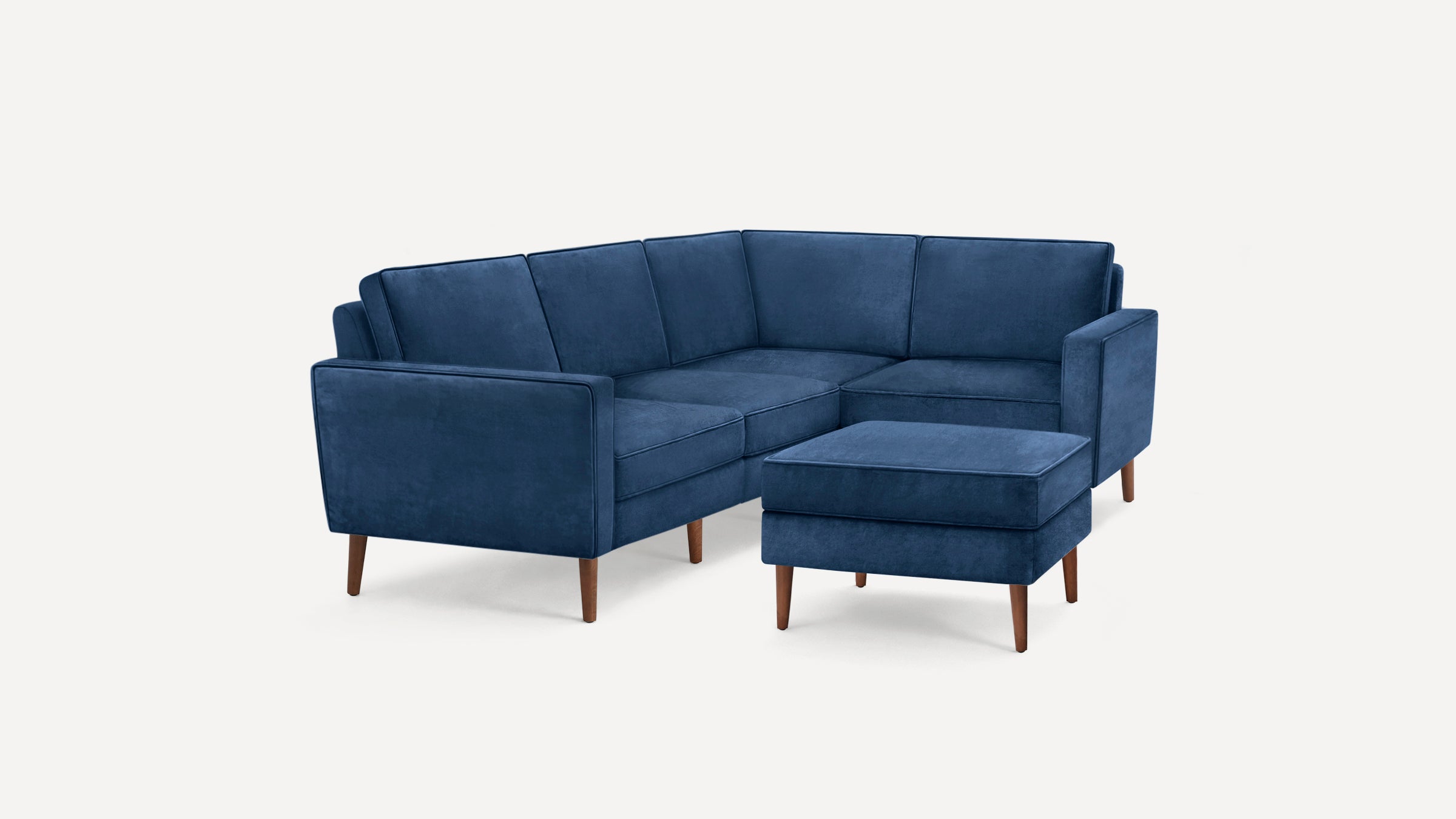 Modern navy blue velvet L-shaped sectional sofa with matching ottoman, supported by tapered wooden legs, set against a white background.