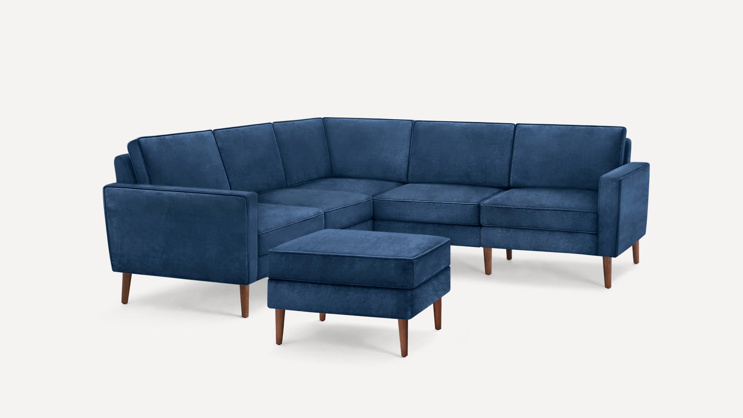 Blue velvet L-shaped sectional sofa with matching ottoman and wooden legs, shown on a white background.
