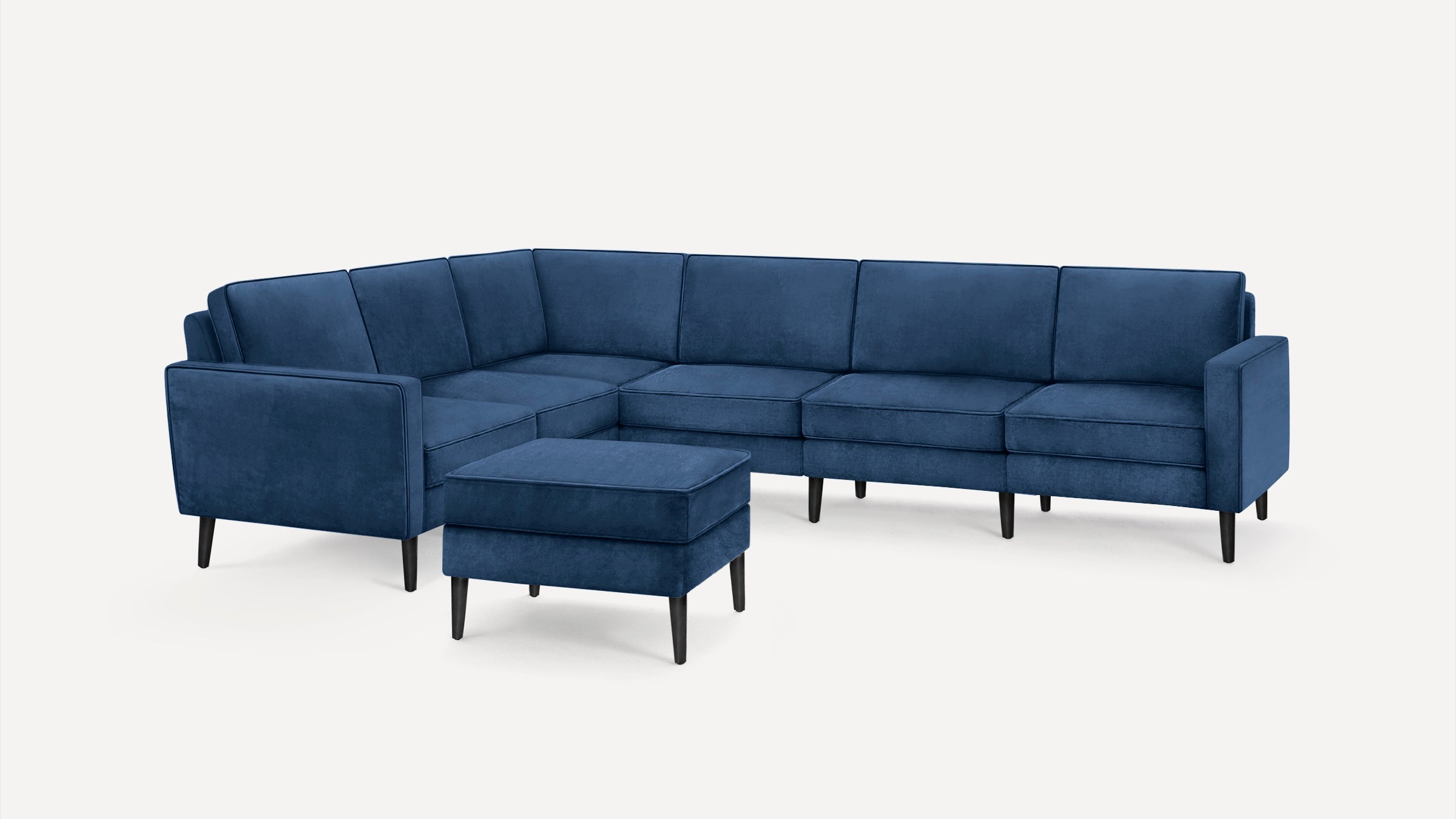 Modern navy blue velvet sectional sofa with angled cushions, black tapered legs, and matching ottoman on white background.