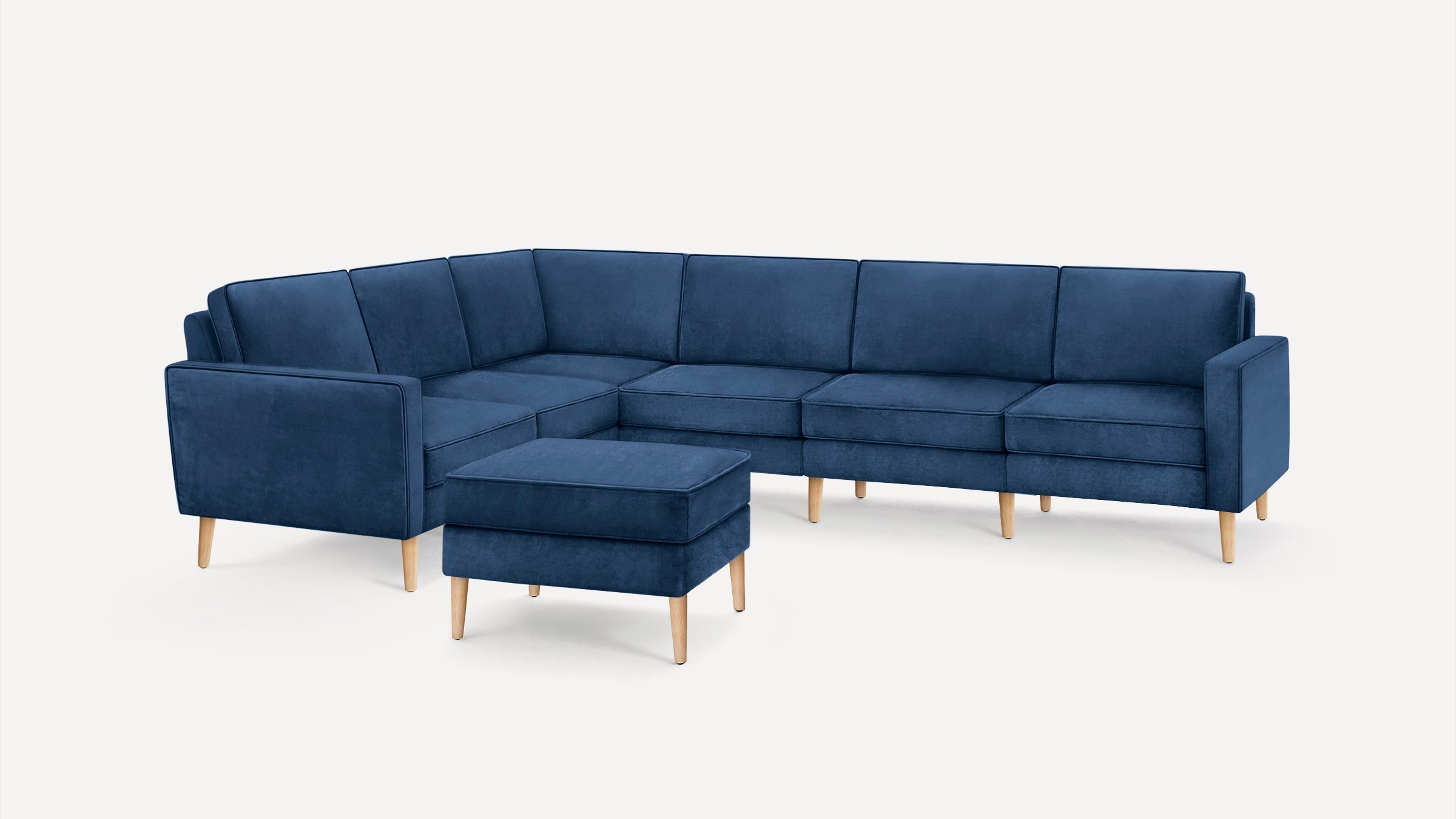 Modern L-shaped sectional sofa in navy blue fabric with light wooden legs, accompanied by a matching ottoman.