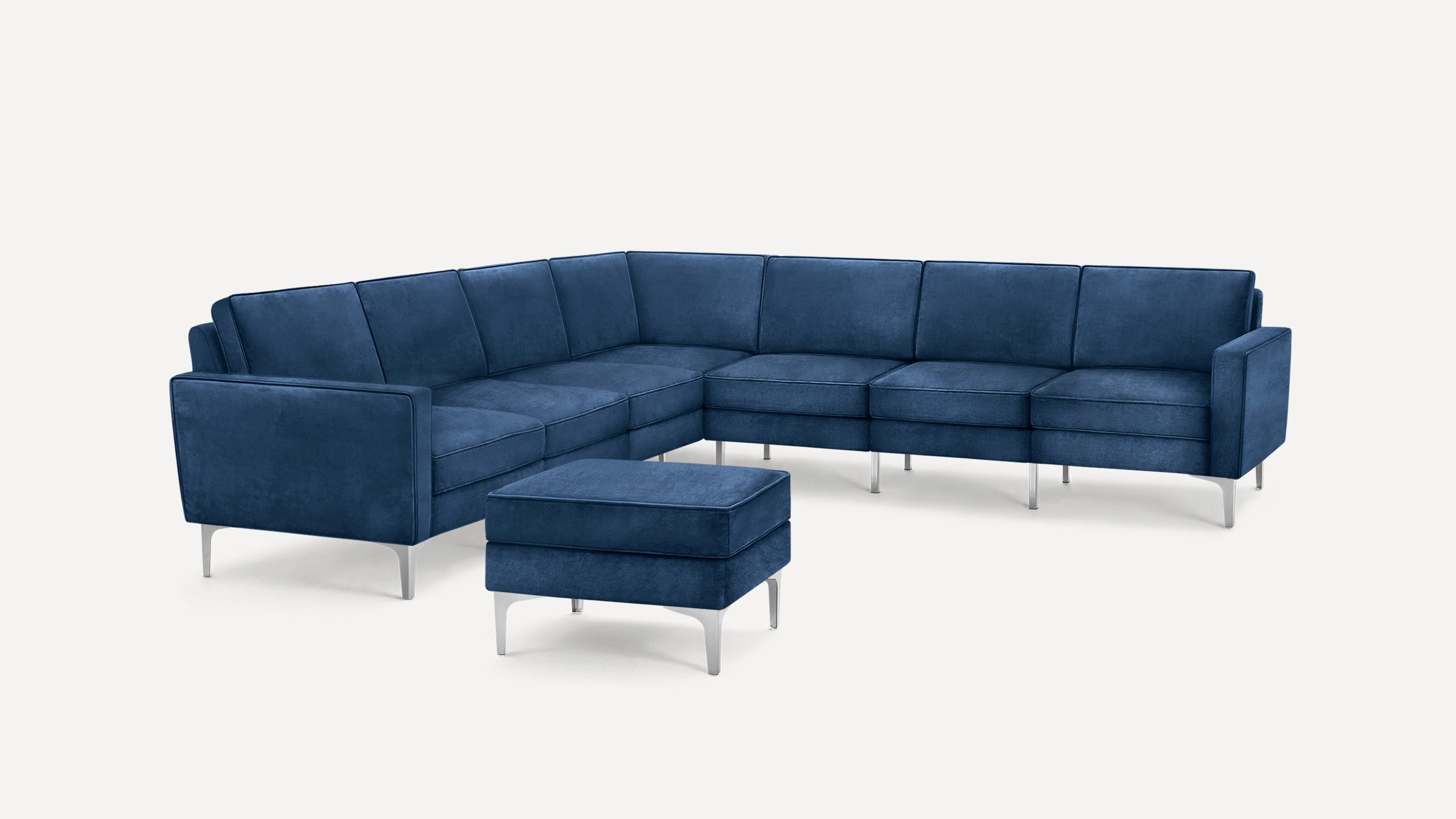 Modern L-shaped sectional sofa with blue velvet upholstery and matching ottoman, featuring sleek silver metal legs.