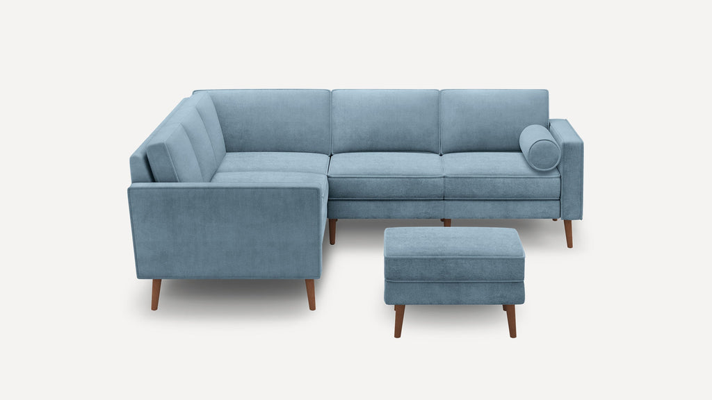Light blue velvet L-shaped sectional sofa with wooden legs and matching ottoman, featuring a cylindrical bolster pillow.