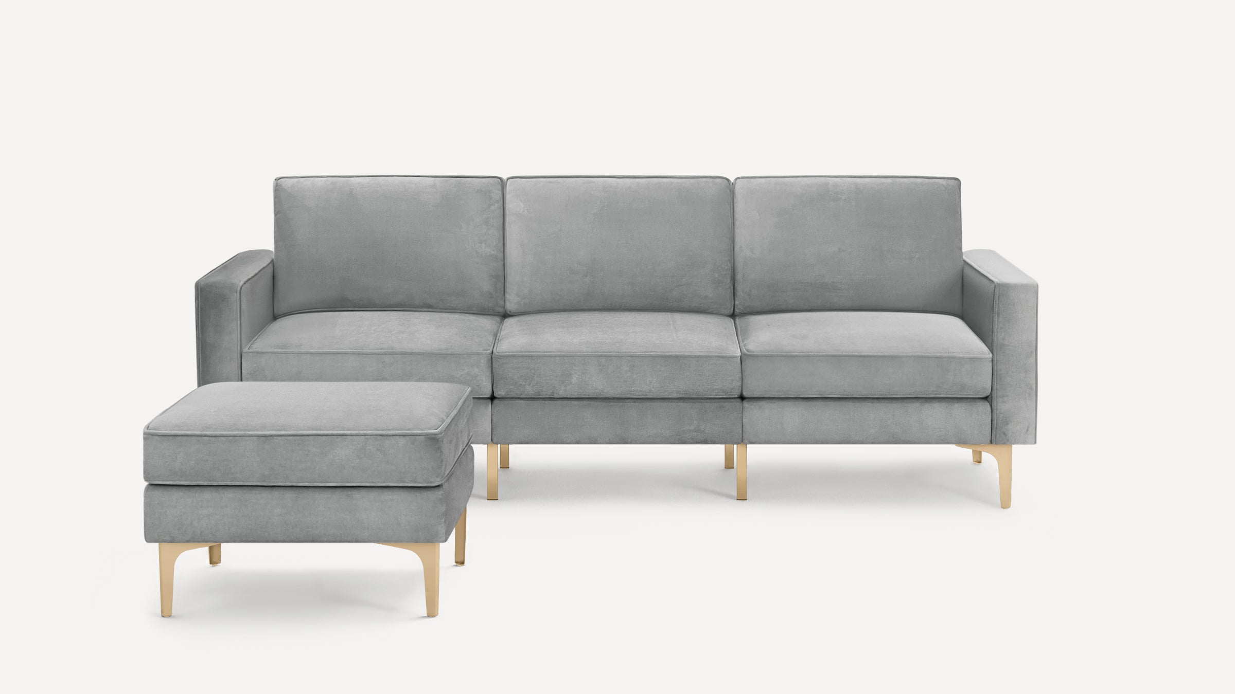 Modern gray velvet three-seater sofa with matching ottoman, both featuring light wooden legs, set on a white background.