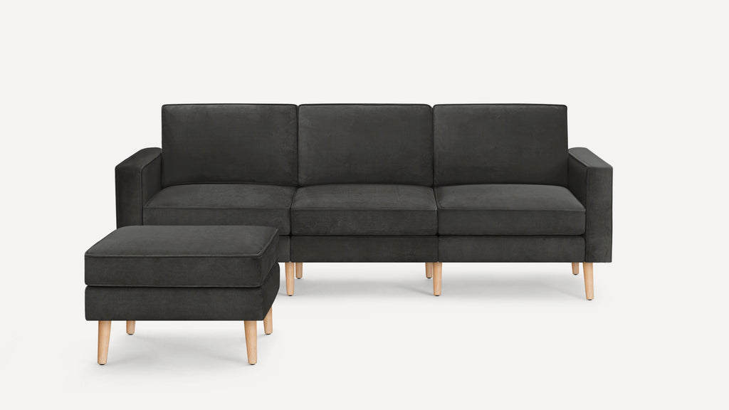 Dark gray mid-century modern three-seater sofa with wooden legs and matching ottoman on a white background.
