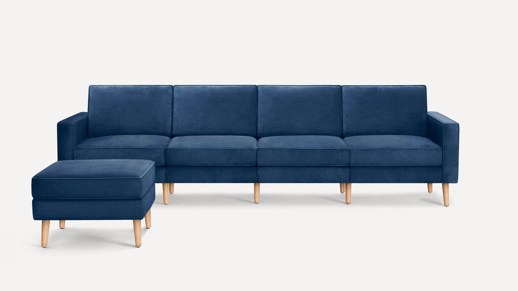 Modern navy blue velvet sofa with matching ottoman, featuring clean lines and light wooden legs, on white background.