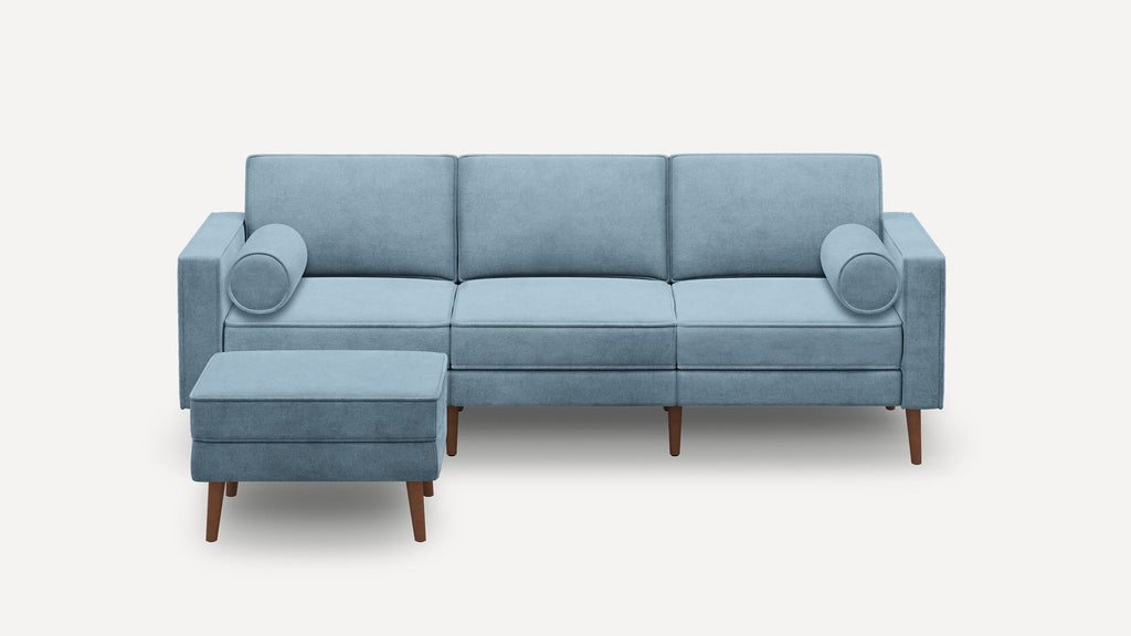 Modern light blue three-seater sofa with matching ottoman, cylindrical bolster pillows, and wooden tapered legs against white background.