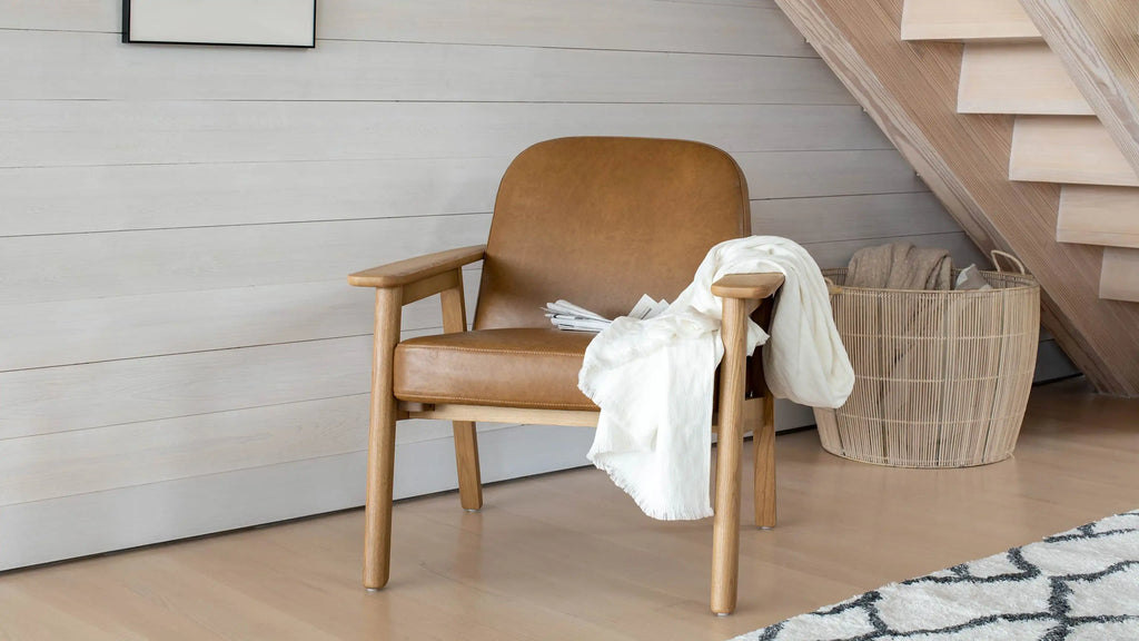 Wood-framed armchair with brown leather cushion, white throw draped over arm, and wicker basket under wooden staircase.