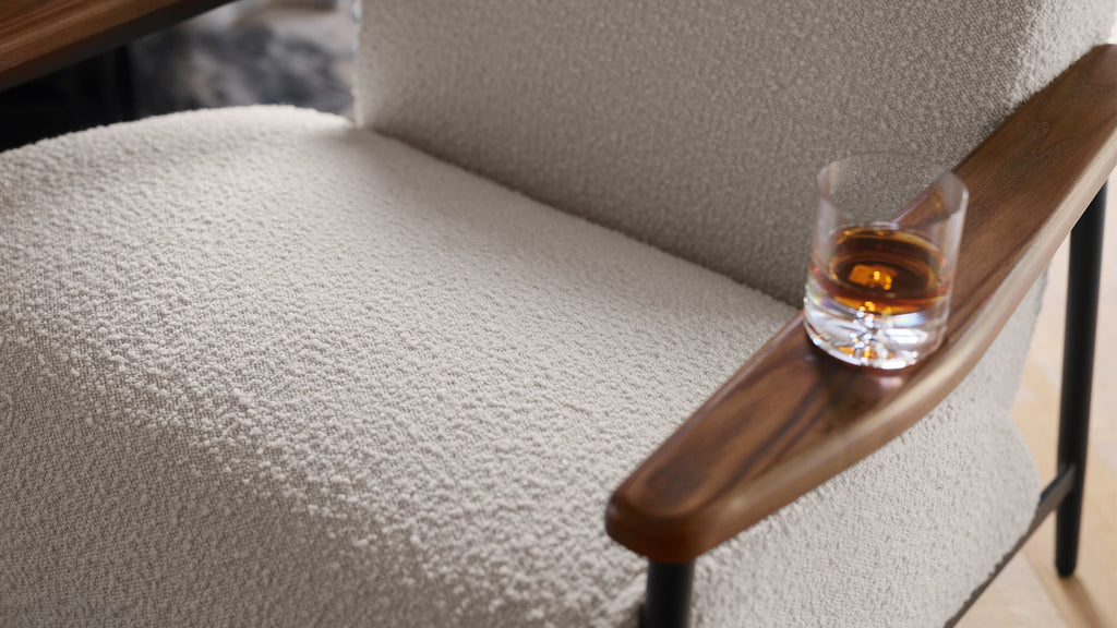 Close-up of a textured beige upholstered chair with a wooden armrest holding a glass of amber liquid.
