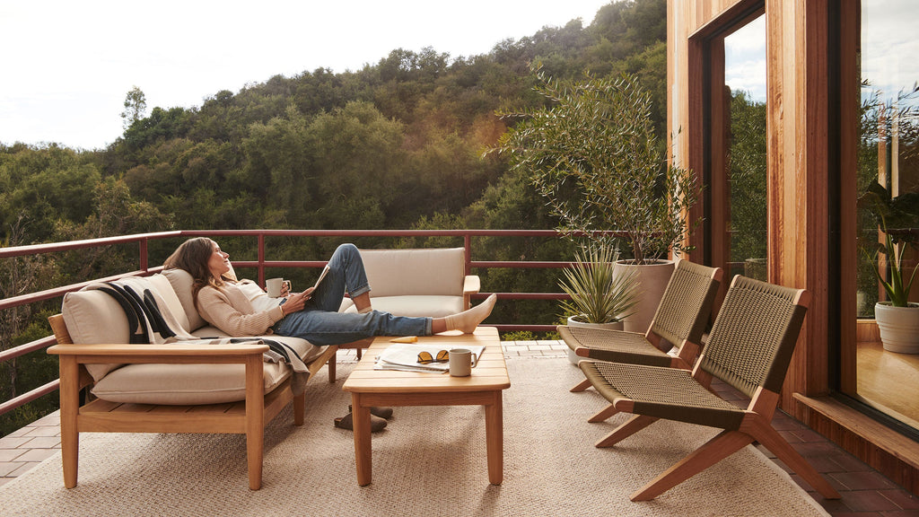 Outdoor wooden patio set with cushioned sofa, two woven chairs, and rectangular wooden coffee table on balcony overlooking green hillside.
