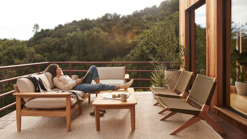 Outdoor patio setup featuring wooden framed cushioned sofa, wooden coffee table, woven chairs, plants, and a person relaxing with coffee and book.