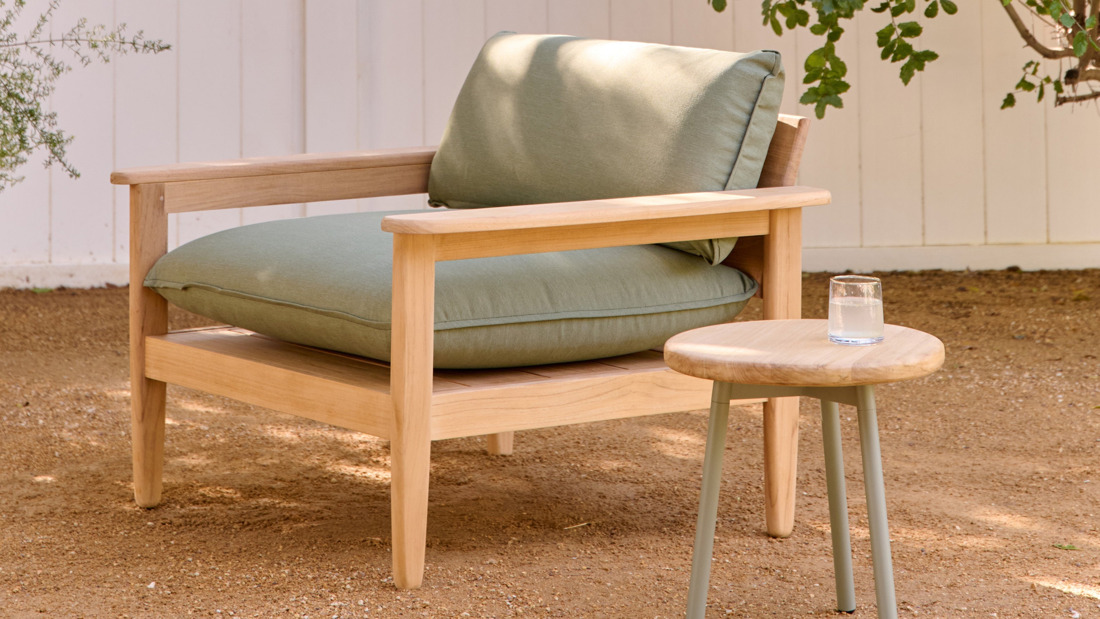 Wooden outdoor armchair with thick sage green cushions next to a small round wooden stool holding a glass of water.