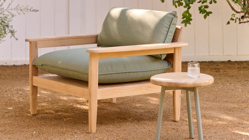 Wooden outdoor armchair with thick sage green cushions next to a small round wooden stool holding a glass of water.