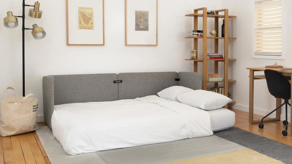 Modern gray fabric fold-out sofa bed in a bright, minimalist living room with wooden floors, bookshelf, and desk.