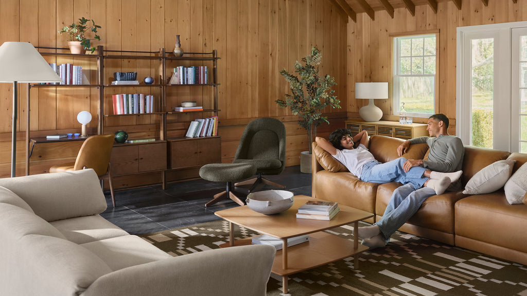 Modern cozy living room with leather sofa, beige couch, green armchair, wooden coffee table, bookshelf, and two people relaxing and smiling.