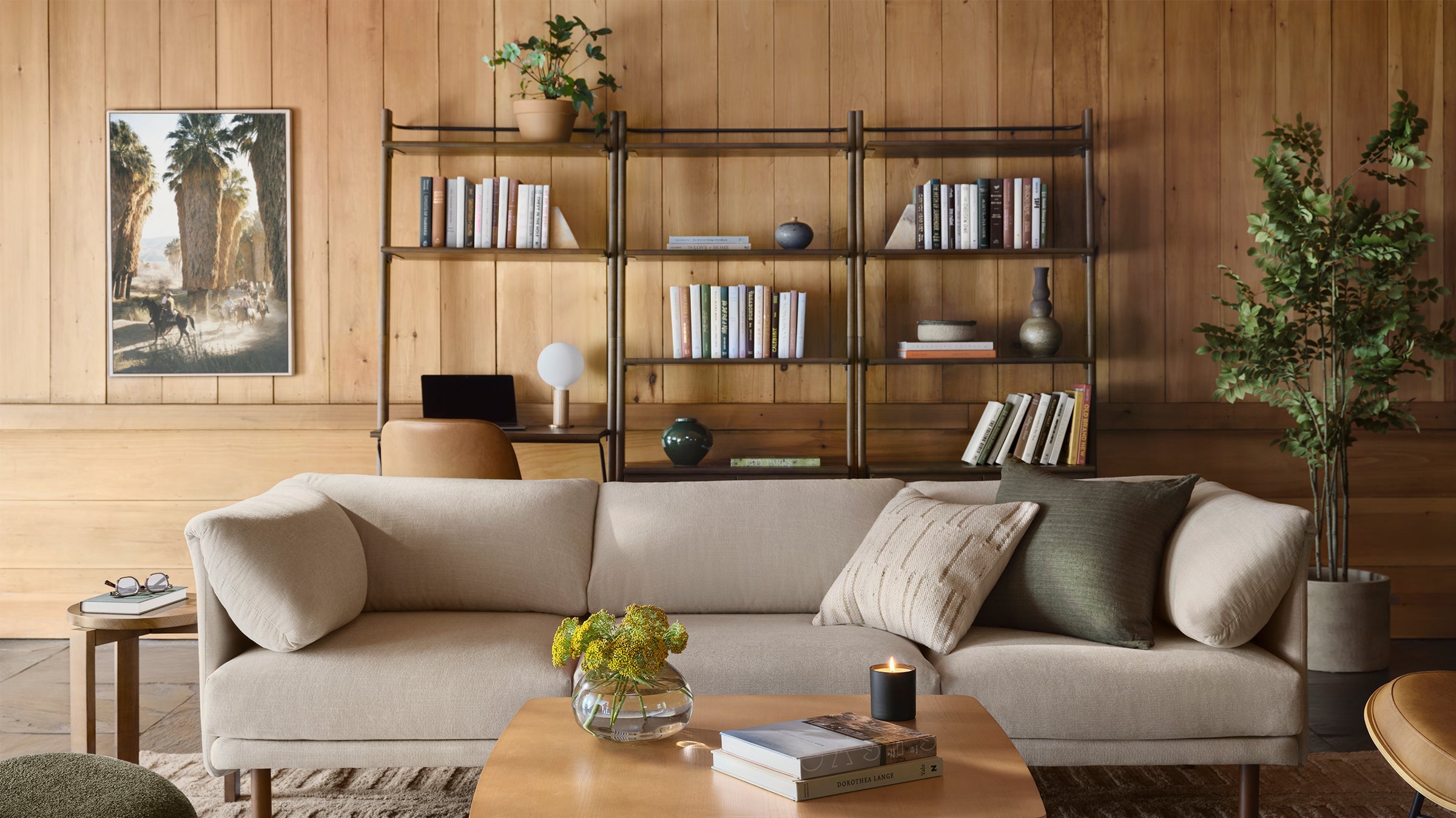 Beige linen sectional sofa with textured pillows in a cozy living room featuring wooden walls, bookshelves, plants, and a coffee table.