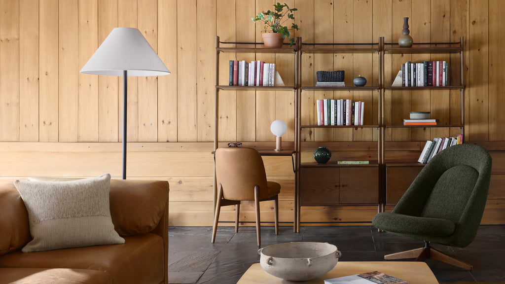 Cozy living room with tan leather sofa, gray floor lamp, wooden bookshelf, olive green swivel chair, and minimalist decor.