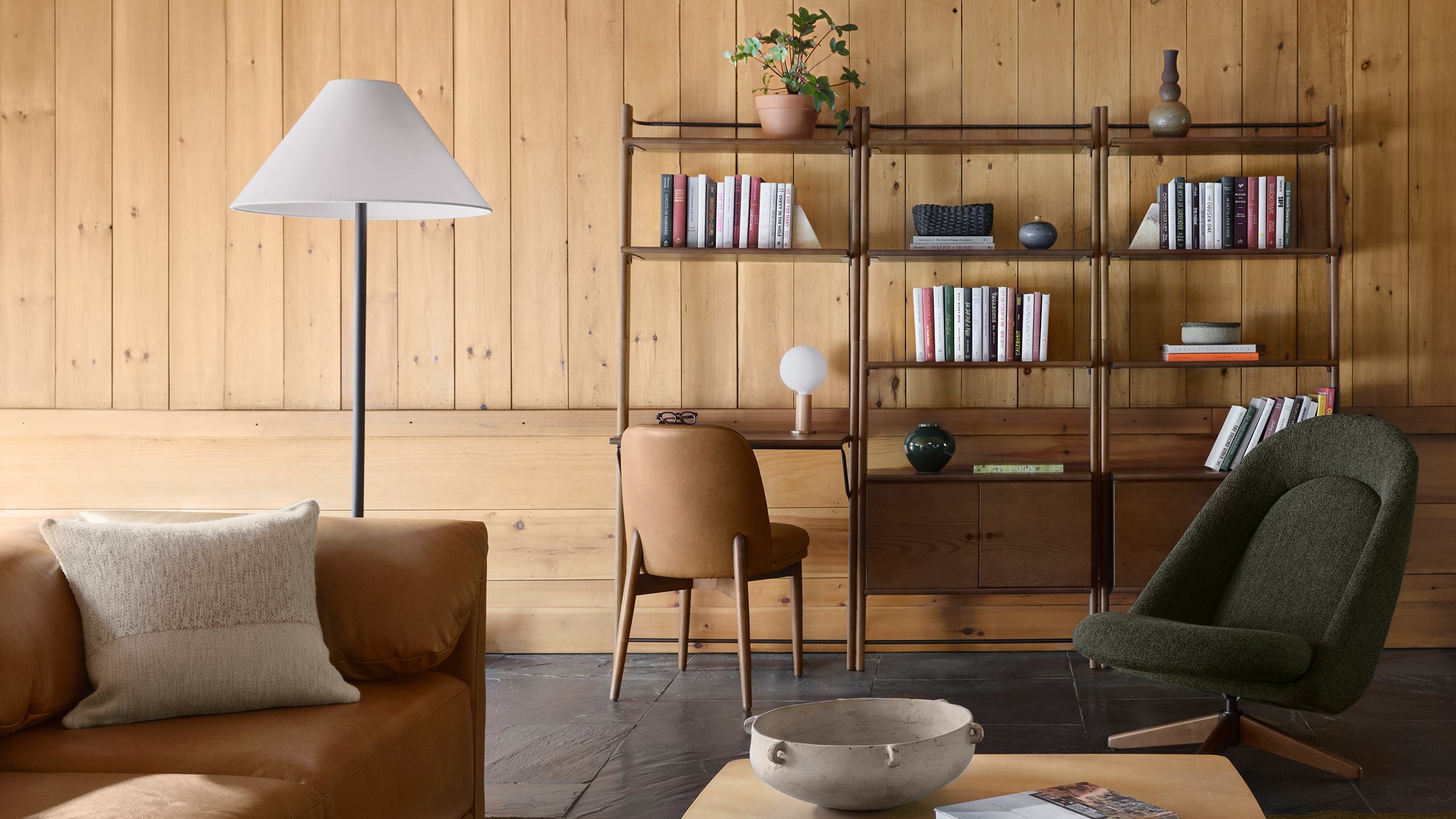 Cozy living room with tan leather sofa, gray floor lamp, wooden bookshelf, olive green swivel chair, and minimalist decor.