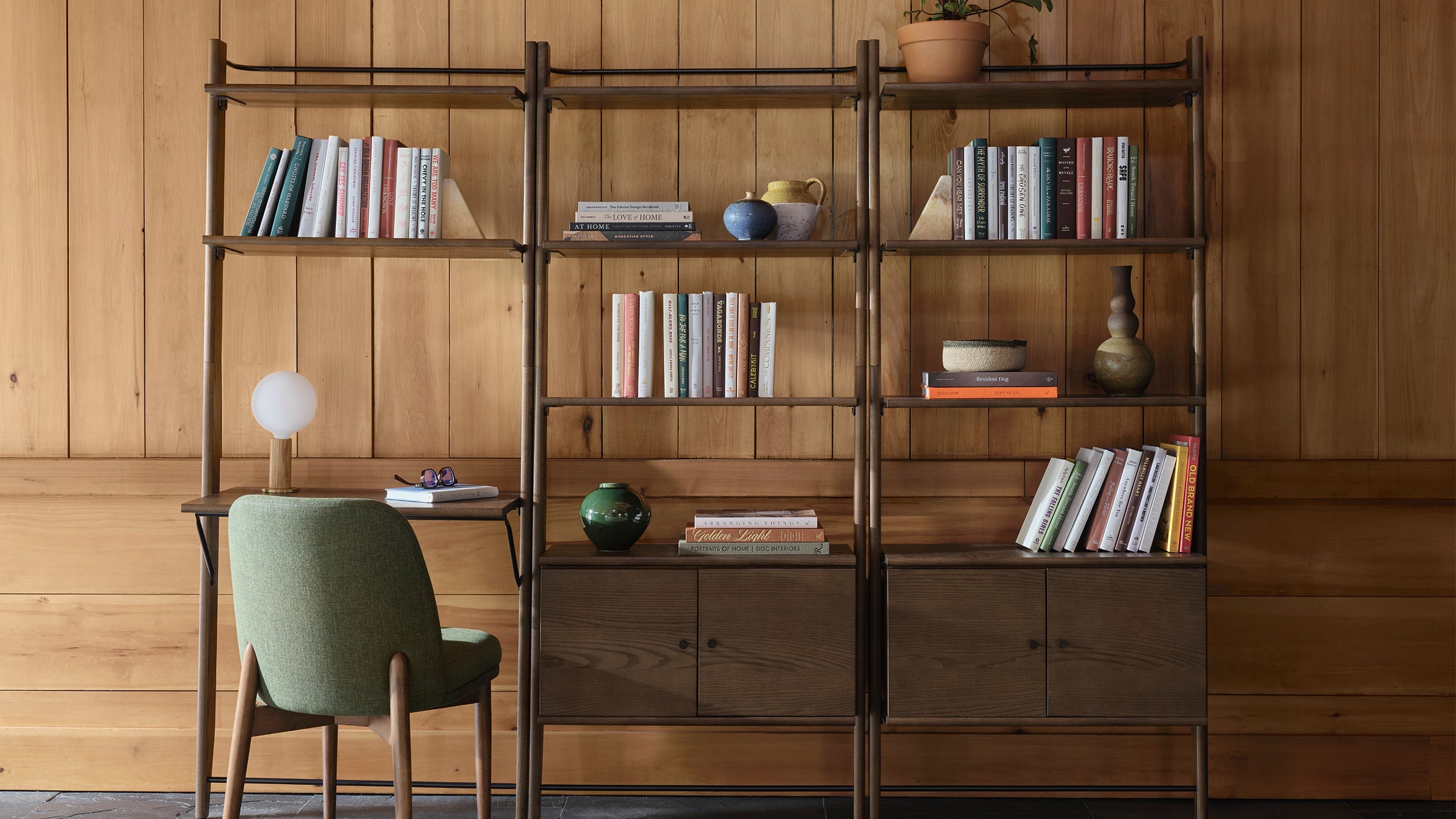 Wooden shelving unit with books, ceramic vases, plant, and attached desk featuring a green upholstered chair and modern table lamp.