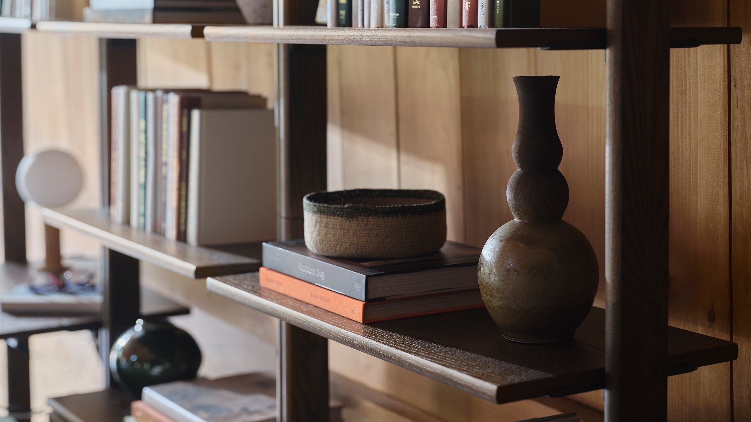 Close-up of wooden shelves with books, a woven basket, and ceramic vases against a wood-paneled wall in soft natural light.