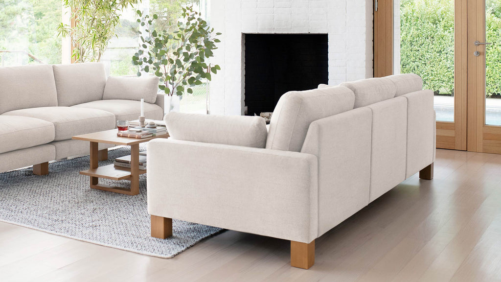 Modern living room with two beige fabric sofas, wooden legs, a wooden coffee table, large rug, fireplace, and large windows.