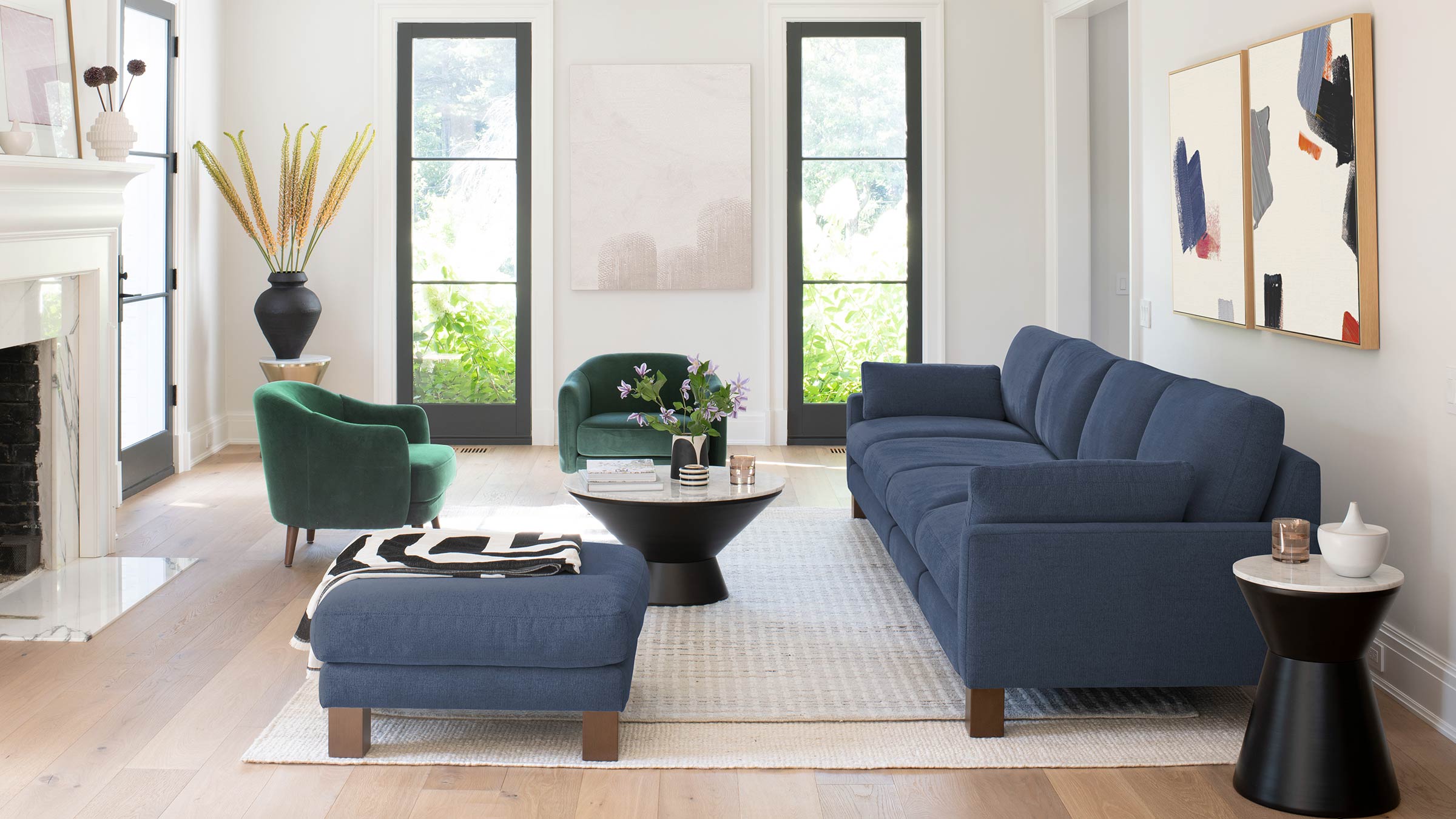 Modern living room featuring a navy blue sofa and ottoman, two green armchairs, black and white coffee table, and abstract wall art.