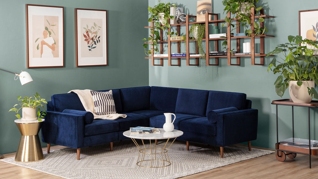 Navy blue velvet L-shaped sectional sofa with wooden legs in a modern living room featuring plants and framed artwork.